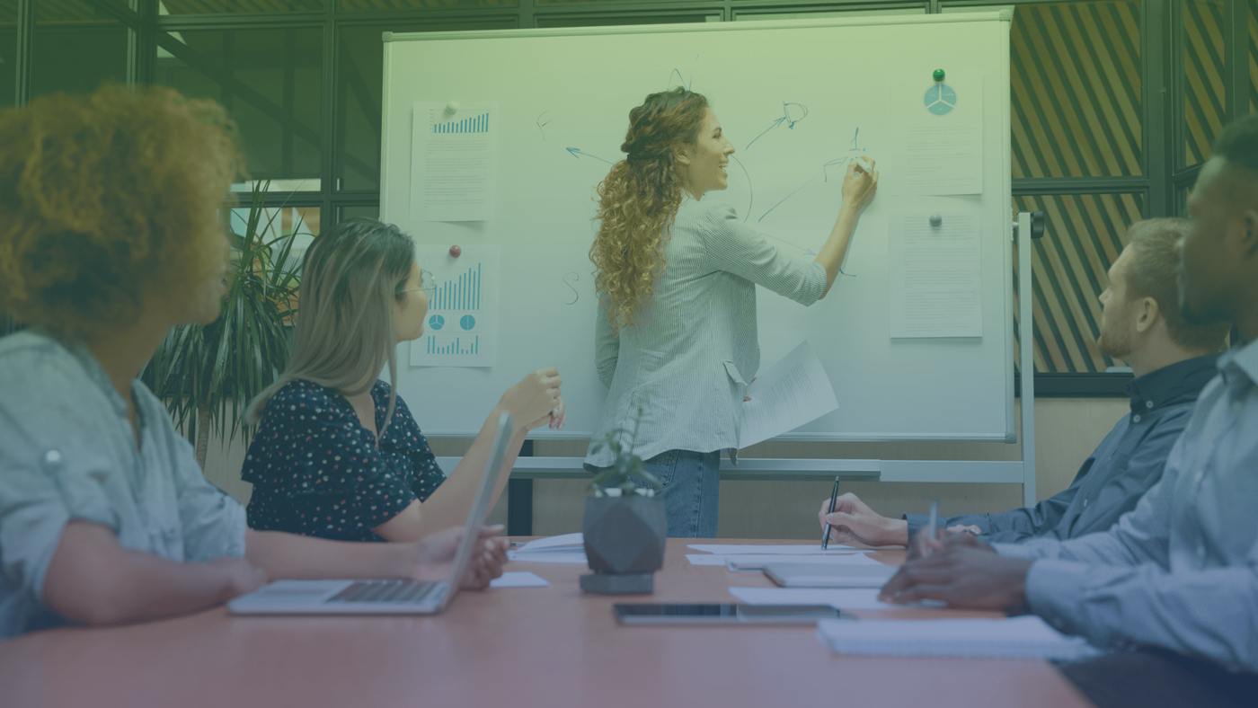 Eine Frau schriebt in einem Meeting am Ende eines Tisches auf einem Whiteboard. Am Tisch sitzen vier Personen und sehen ihr zu.