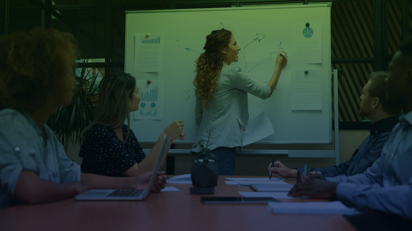 Eine Frau schriebt in einem Meeting am Ende eines Tisches auf einem Whiteboard. Am Tisch sitzen vier Personen und sehen ihr zu.