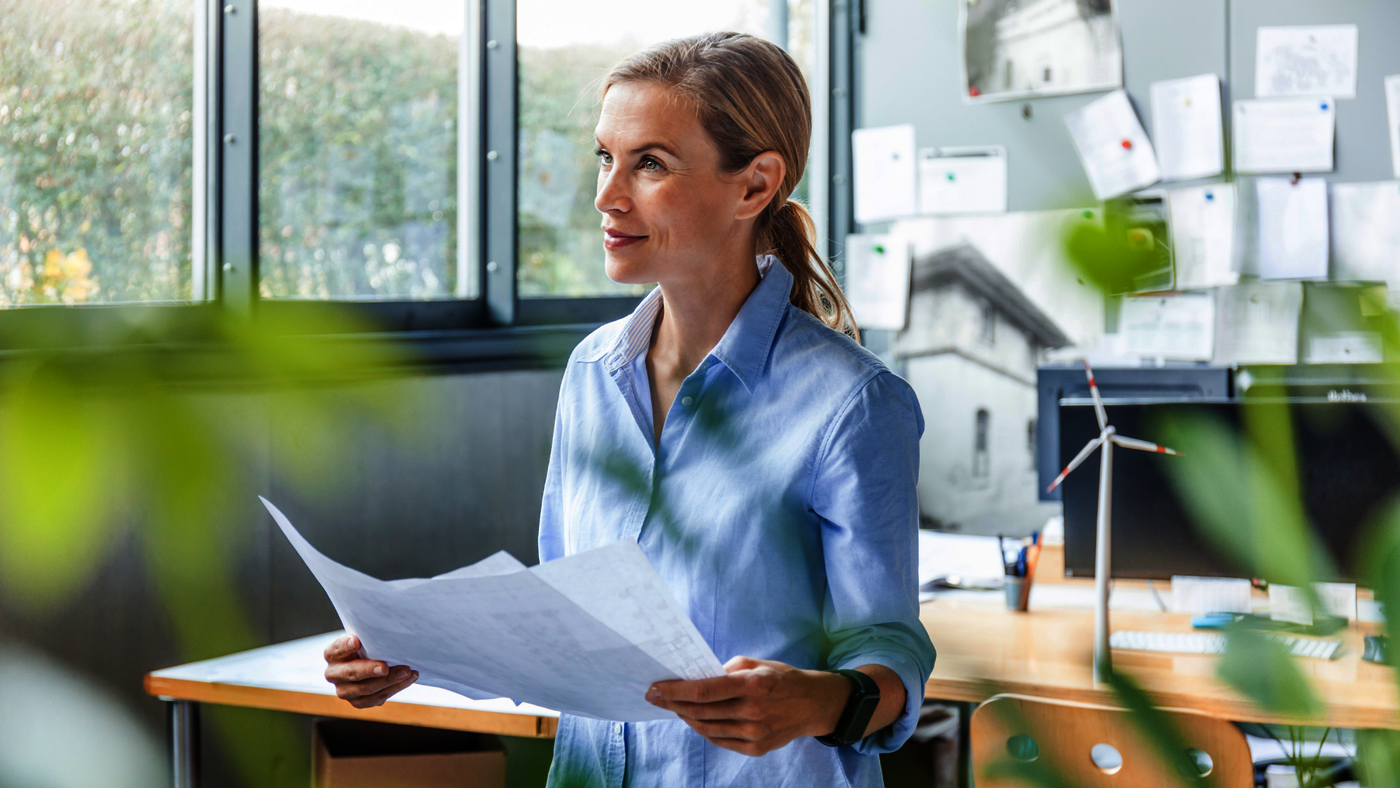 Eine Architektin steht in ihrem Büro und hält ein Blatt Papier, wahrscheinlich einen Plan, in ihrer Hand.