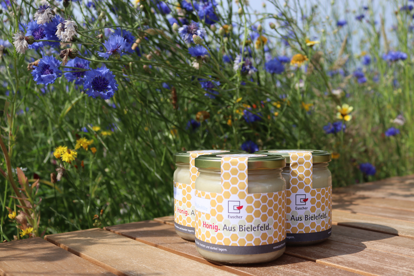 Das Bild zeigt einen Blumenwiese und davor den eigenen Honig der Euscher GmbH & Co. KG