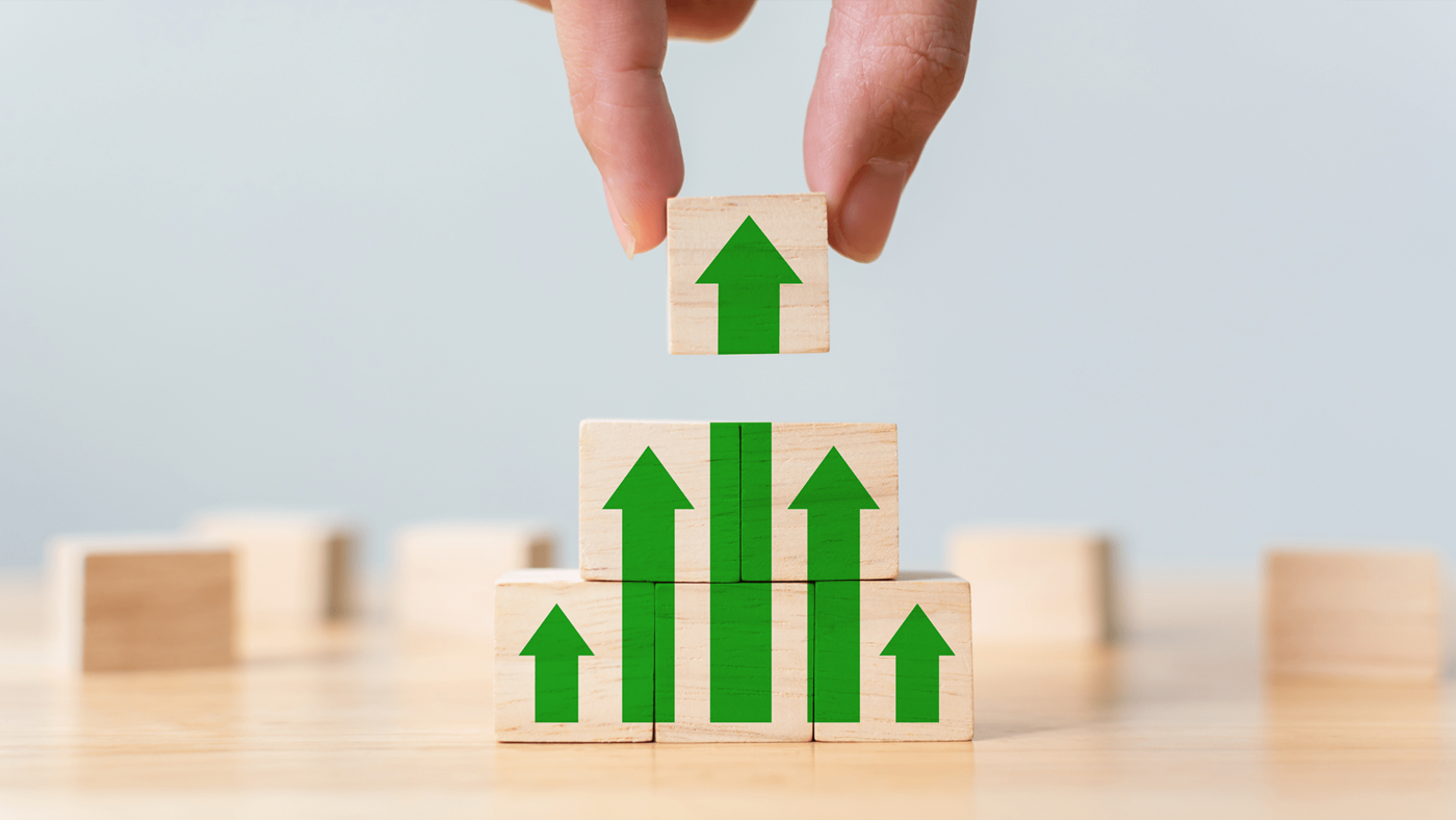 Eine Hand stapelt Bausteine mit grünen Markierungen übereinander, die zusammen nach oben zeigende Pfeile ergeben.