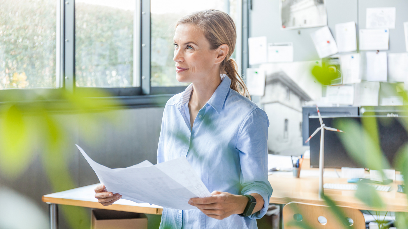 Eine Architektin steht in ihrem Büro und hält ein Blatt Papier, wahrscheinlich einen Plan, in ihrer Hand.